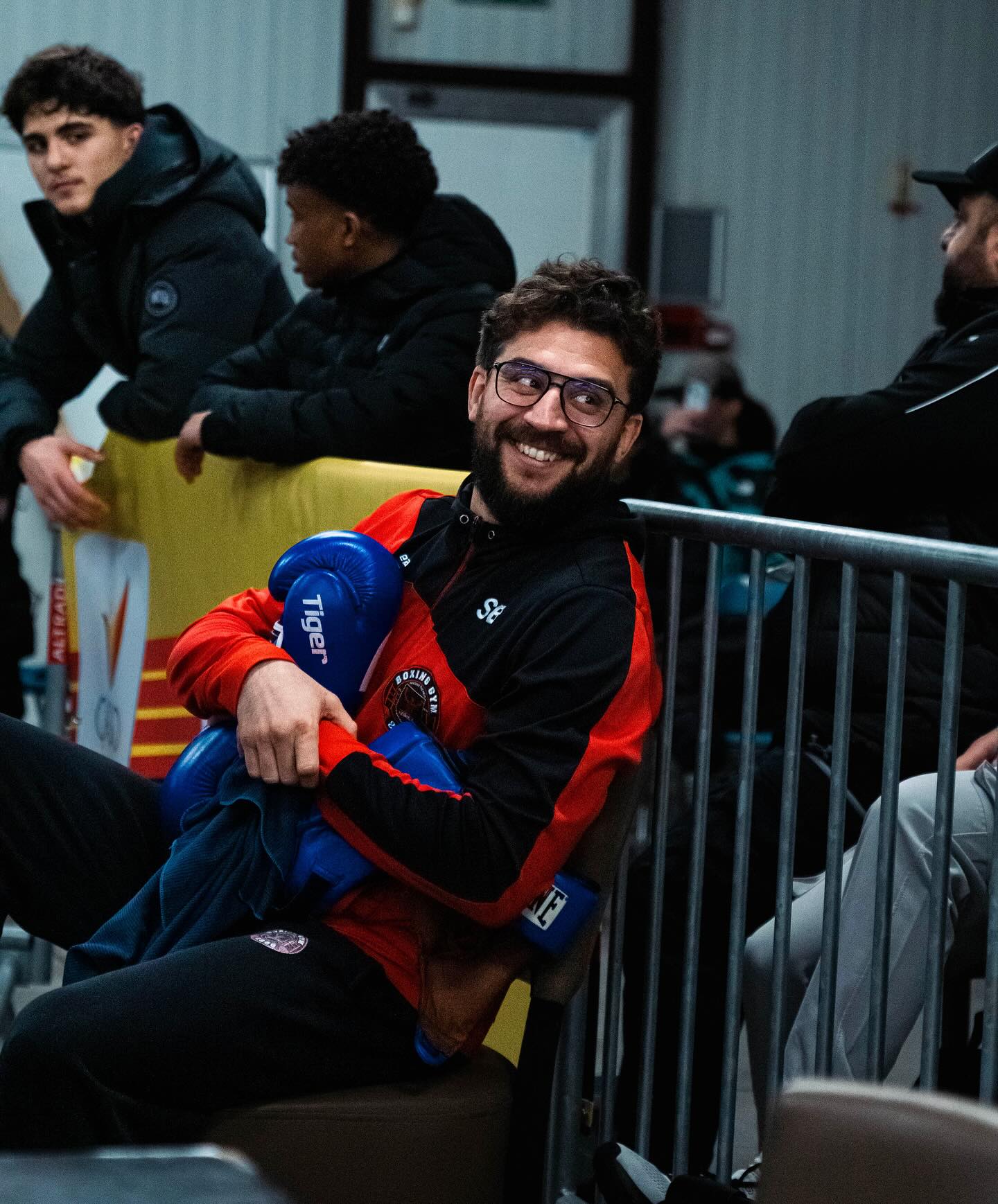 Een foto van een lachende man die bokshandschoenen vastheeft en een rood vest draagt van Boxing Gym 2800. Deman is bekend als Sabri Belhadj en hij is de oprichter van Boxing Gym 2800, 015 Boxing en C-Suite Boxing
